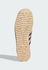 Světlé podrážka tenisek s texturovanou gumou, kruhovými vzory trakce a třemi hnědými pruhy; na boku je značka Adidas.