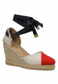 Espadrille compensée avec un dessus en toile rouge, beige et blanc. Présente des détails en fibres naturelles tissées au talon et des liens noirs à la cheville.