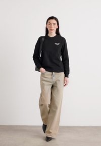 En person går selvsikkert i en svart hettegenser og lysebeige bukser, kombinert med svarte hæler, mot en enkel hvit bakgrunn.