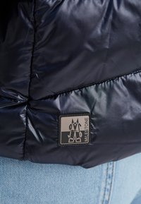 Marineblaue gepolsterte Jacke mit glänzender Textur und quadratischem schwarzen und grauen Logoaufnäher, ausgestattet mit gesteppten Abschnitten und einer glatten Oberfläche.