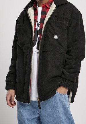 Mężczyzna ubrany w czarną sztruksową kurtkę z polarową podszewką, czerwoną koszulę w kratę, białą koszulkę z nadrukiem i jasnoniebieskie dżinsy, ręka w kieszeni.