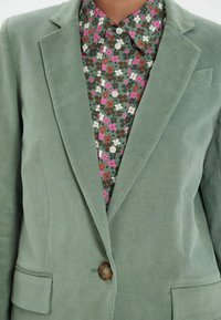 Blazer en velours vert avec deux poches avant et une fermeture à boutons, porté par-dessus une chemise à motif floral dans des teintes de rose et de brun.