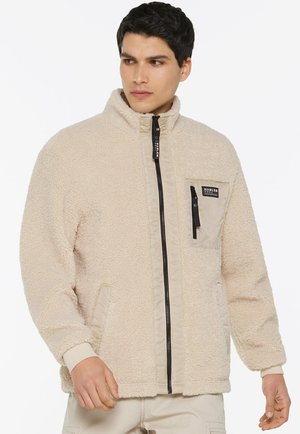 Man wearing beige fleece jacket with front zipper and zippered chest pocket, looking to his left.