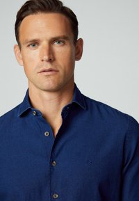 Camicia button-up blu navy in un tessuto morbido con colletto classico. Presenta bottoni marroni e un piccolo logo ricamato sul petto.