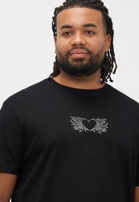 Μαύρο t-shirt με κοντά μανίκια κατασκευασμένο από βαμβάκι. Διαθέτει λευκό γραφικό σχέδιο καρδιάς με φτερά που μοιάζουν με φλόγες στο στήθος.