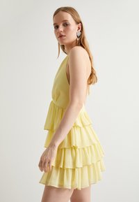 Robe sans manches jaune à volants avec un corsage ajusté et un ourlet en étages. Fabriquée en tissu doux et léger avec une texture lisse.