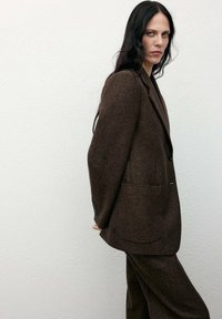 Blazer in lana marrone con rever a notch, chiusura a un bottone e due tasche frontali, abbinato a pantaloni coordinati. Tessuto testurizzato, vestibilità su misura.