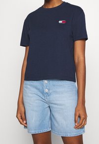 Osoba v námořnickém modrém tričku Tommy Jeans a světlých denimových šortkách s třemi předními knoflíky na jednobarevném pozadí.