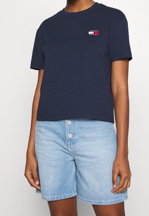 Osoba v námořnickém modrém tričku Tommy Jeans a světlých denimových šortkách s třemi předními knoflíky na jednobarevném pozadí.