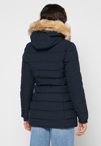 Giacca imbottita blu navy con motivo trapuntato orizzontale, cappuccio e rifinitura in pelliccia sintetica. Presenta una silhouette aderente per calore e comfort.