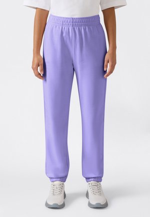 Persoon draagt lila joggers met elastische taille en witte sneakers, staand tegen een effen witte achtergrond.