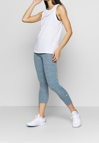 Biała top bez rękawów, niebieskie melanzowe legginsy i białe buty sportowe. Legginsy mają logo Nike po lewej stronie. Gładki, casualowy design.