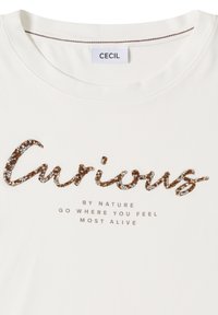 T-shirt blanc avec le mot « Curious » en strass marron et argent, et un texte plus petit disant « Par nature, va là où tu te sens le plus vivant. »