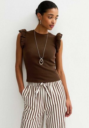 Femme aux cheveux foncés attachés en chignon portant un haut marron sans manches avec des épaules à volants, un pantalon rayé blanc et marron, et un long collier en argent.
