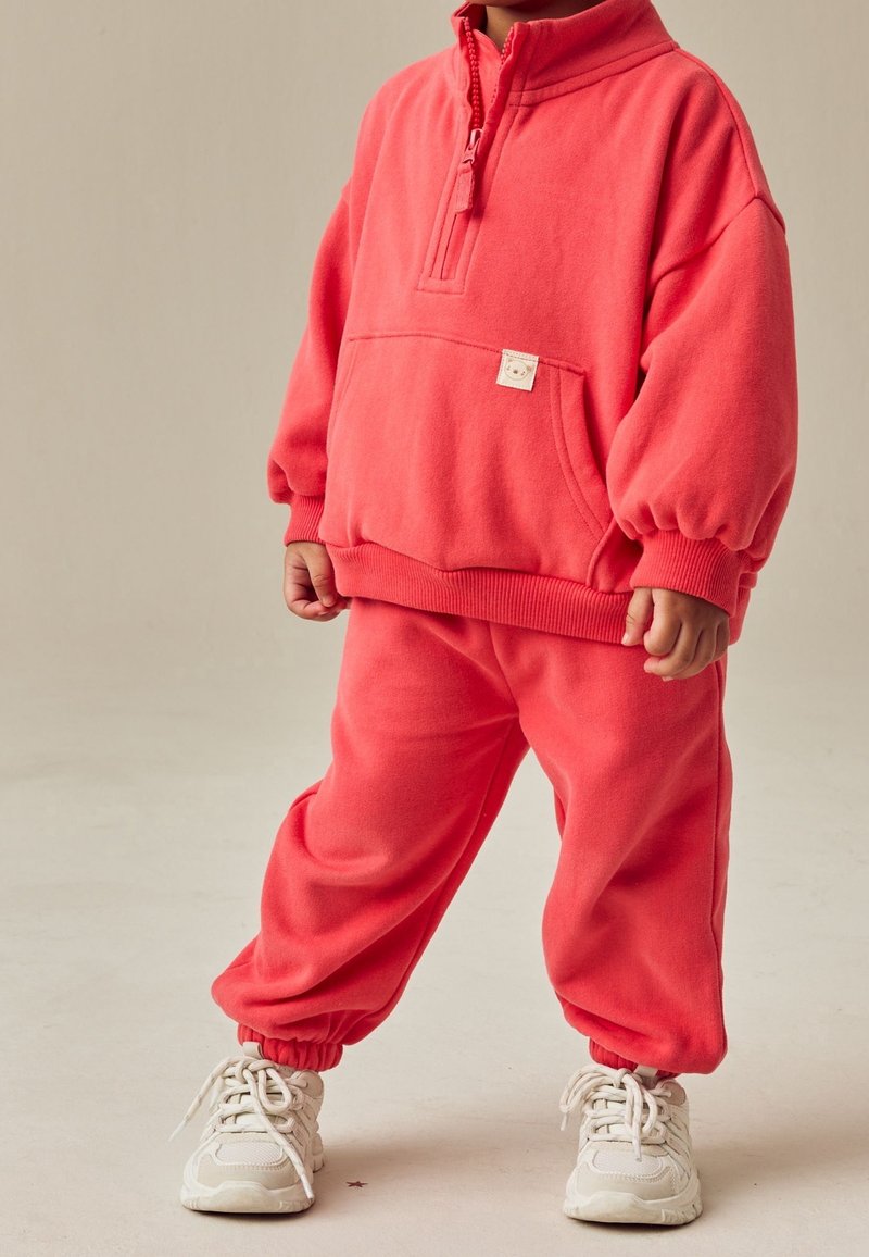 Enfant portant un sweat-shirt en polaire corail vif et un pantalon assorti, avec des baskets blanches, debout devant un fond beige uni.