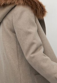 Abrigo de lana beige con cuello de piel marrón, usado sobre un suéter gris, mostrando el brazo izquierdo y el hombro en una vista en primer plano.