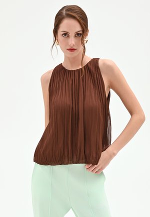 Donna con top marrone plissettato senza maniche e pantaloni verde chiaro, che indossa orecchini geometrici dorati, posando contro uno sfondo bianco uniforme.