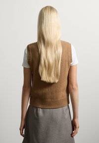 Brown knitted sleeveless sweater with ribbed edges, worn over a white shirt. Long, straight blonde hair cascades down the back.