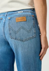 Jeans de mezclilla azul con un parche de cuero marrón que dice "Wrangler" y costuras en zigzag distintivas en los bolsillos traseros.