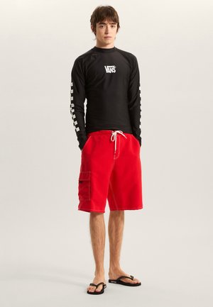 Sort langærmet rashguard med hvidt Vans-logo og ternet mønster på ærmerne; røde cargo badeshorts; sorte flip-flops.