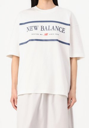 Osoba nosząca luźną, białą koszulkę New Balance z granatowymi paskami i napisem, w połączeniu z jasnymi spodniami lub spódnicą.