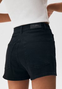 Černé džínové šortky s vysokým pasem, dvěma zadními kapsami a texturovanou látkou. Na pásku je štítek značky „PULL&BEAR“.