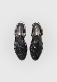 Juodos odinės šlepetės su pintomis dirželiais, atvirais pirštais ir slingback dizainu, kuriame yra reguliuojama sagtis ant kulkšnies. Plokščias padas.