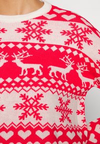 Röd och vit stickad tröja med ett mönster av renar och snöflingor, med en ribbad halsringning och öppet armhålsområde för ventilation.