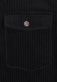 Tissu en velours côtelé noir avec des côtes verticales, présentant une poche sur la poitrine avec fermeture à bouton et de petits détails de couture.