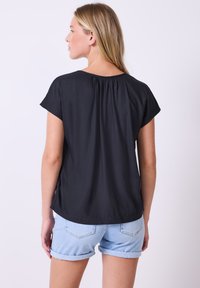 Donna con capelli biondi che indossa una maglietta nera a maniche corte e larga e shorts di jeans azzurri arrotolati, girata di spalle su uno sfondo semplice.