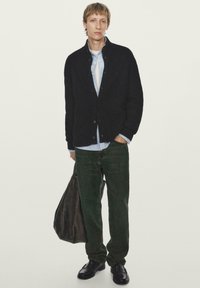 Černý žebrovaný cardigan, modrá košile na knoflíky, tmavě zelené džíny, černé boty a hnědá kožená taška. Jednoduchý design s neutrálními tóny.