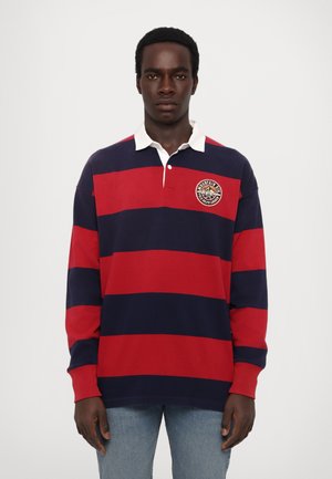 Rød og marineblå stribet rugbyshirt med lange ærmer, en hvid krave og et rundt logo-patch på venstre bryst. Fremstillet i bomuldsmateriale.