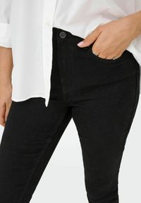 Mustad kõrgpuusadega teksad, millel on ülikitsas vorm, näidates sujuvat tekstuuri, esimesi taskuid ja metallist nööbist sulgemist vööl.