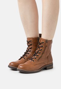 Bottes chevilles en cuir marron avec lacets, une fermeture éclair latérale, un bout rond et un petit talon. Présente des détails de coutures et des accents en métal.