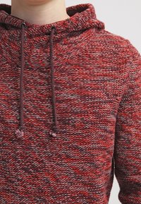 Rote und graue Strickjacke mit Stehkragen, verstellbaren Kordeln und einem strukturierten Muster. Warmes und lässiges Design, das sich gut zum Schichten eignet.