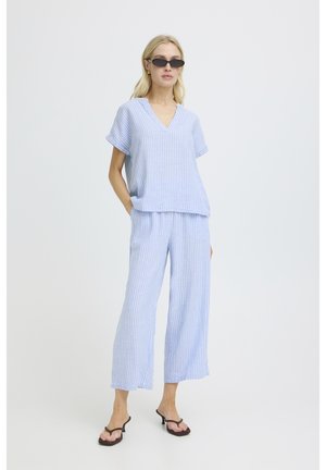 Sieviete valkā gaiši zilu un baltu svītrainu īsroku topu un platajiem biksēm, melnas saulesbrilles ar kaķa acu formu, melnas papēžu sandales, stāv ar roku kabatā.