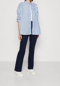 Camisa de botones a rayas azules y blancas con mangas largas, combinada con jeans acampanados de color azul oscuro y zapatillas blancas sin cordones.