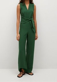 Jumpsuit vert sans manches avec un décolleté en V, détail de taille à nouer, jambes larges et tissu texturé. Porté avec des sandales noires à lanières.