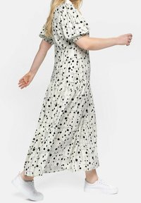 Weißes Kleid mit schwarzen geometrischen Mustern, Puffärmeln und gestuftem Saum; kombiniert mit weißen Sneakers, in einer Seitenansicht.