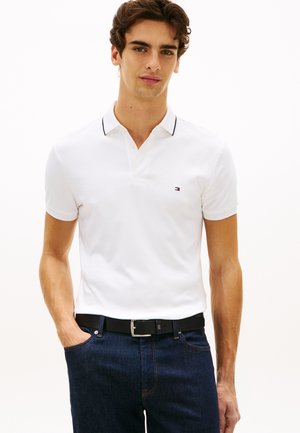 Junger Mann trägt ein weißes Polo-Shirt mit kurzen Ärmeln und kleinem Logo auf der Brust sowie dunkelblaue Jeans, steht mit einer Hand in der Tasche.