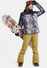 Snowboarder portant une veste à motifs violets et gris avec des accents noirs, un pantalon de neige jaune et des bottes noires, tenant un snowboard.