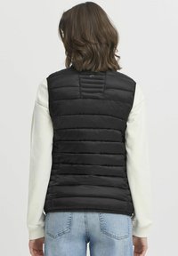 Gilet en doudoune noir avec un design matelassé, un col haut et des détails au dos. Porté sur un haut de couleur crème, associé à un jean bleu clair.