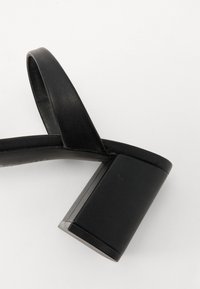 Sandale noire à talons hauts avec un bout arrondi, une large lanière et un talon carré épais, le tout avec une texture lisse et mate.