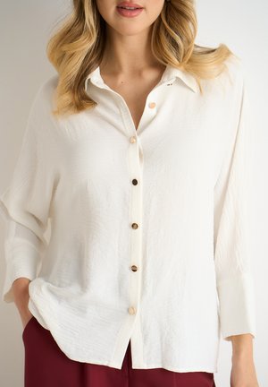 White button-up shirt made of textured fabric with a collar, featuring five buttons and long sleeves. Paired with burgundy pants.