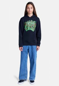 Mladý člověk stojící, oblečený v černé mikině s zelenou grafikou, volných modrých džínách a černých teniskách s bílou podešví, na bílém pozadí.