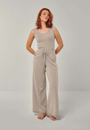 Junge Frau mit roten Haaren steht barfuß, trägt ein beiges ärmelloses Oberteil und weite Hosen mit Kordelzug vor einem einfarbigen grauen Hintergrund.