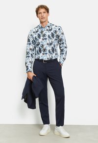 Bloemige overhemd met knopen in blauw en zwarttonen, gecombineerd met marineblauwe broek en witte sneakers. Man houdt een marineblauwe jas over zijn arm.