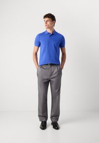 Camisa polo azul de mangas curtas com colarinho, combinada com calças de fato cinzas claras e sapatos pretos, em pé contra um fundo branco liso.