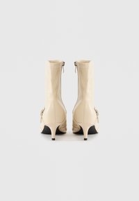 Beige leather ankle boots with side zippers, pointed toes, small kitten heels, and decorative buckled straps, viewed from the back on a white background.
