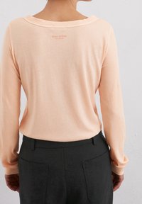 Hellpfirsichfarbener Pullover mit weitem Halsausschnitt, langen Ärmeln und gerippten Bündchen. Schwarze, taillierte Hose mit Rückentaschen. Branding in der Nähe des Kragens.